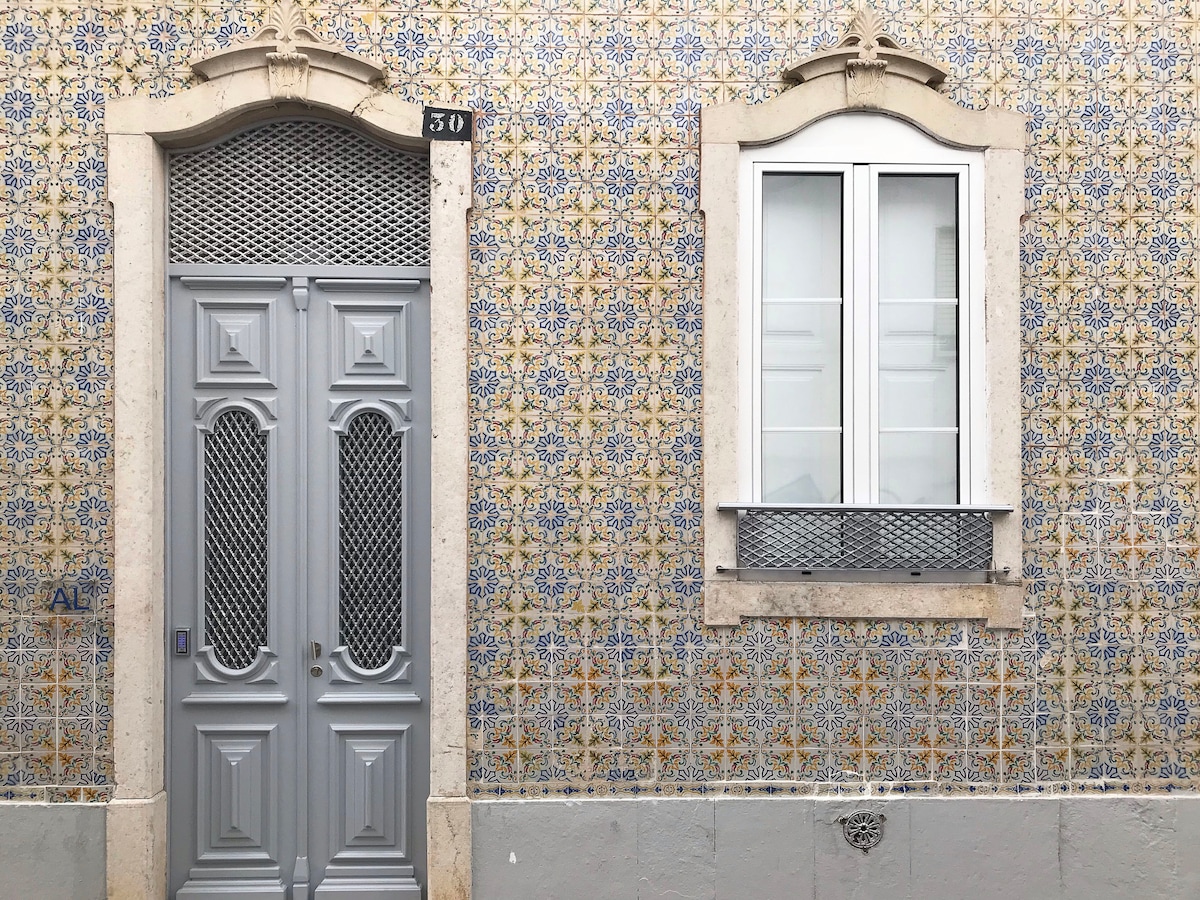 The image features a traditional facade adorned with decorative tiles, showcasing intricate patterns. A grey door with ornate detailing is positioned beside a white window framed in light stone. The wall's vibrant tiles contribute to the historical charm of the building.