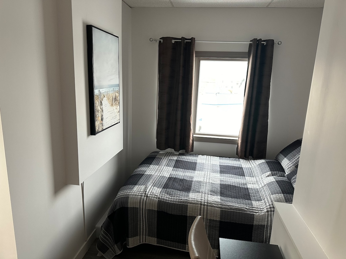 A compact bedroom features a single bed covered in a plaid blanket. Natural light enters through a window adorned with dark curtains, illuminating the room's neutral walls. A simple desk is positioned against one wall, complementing the inviting atmosphere.