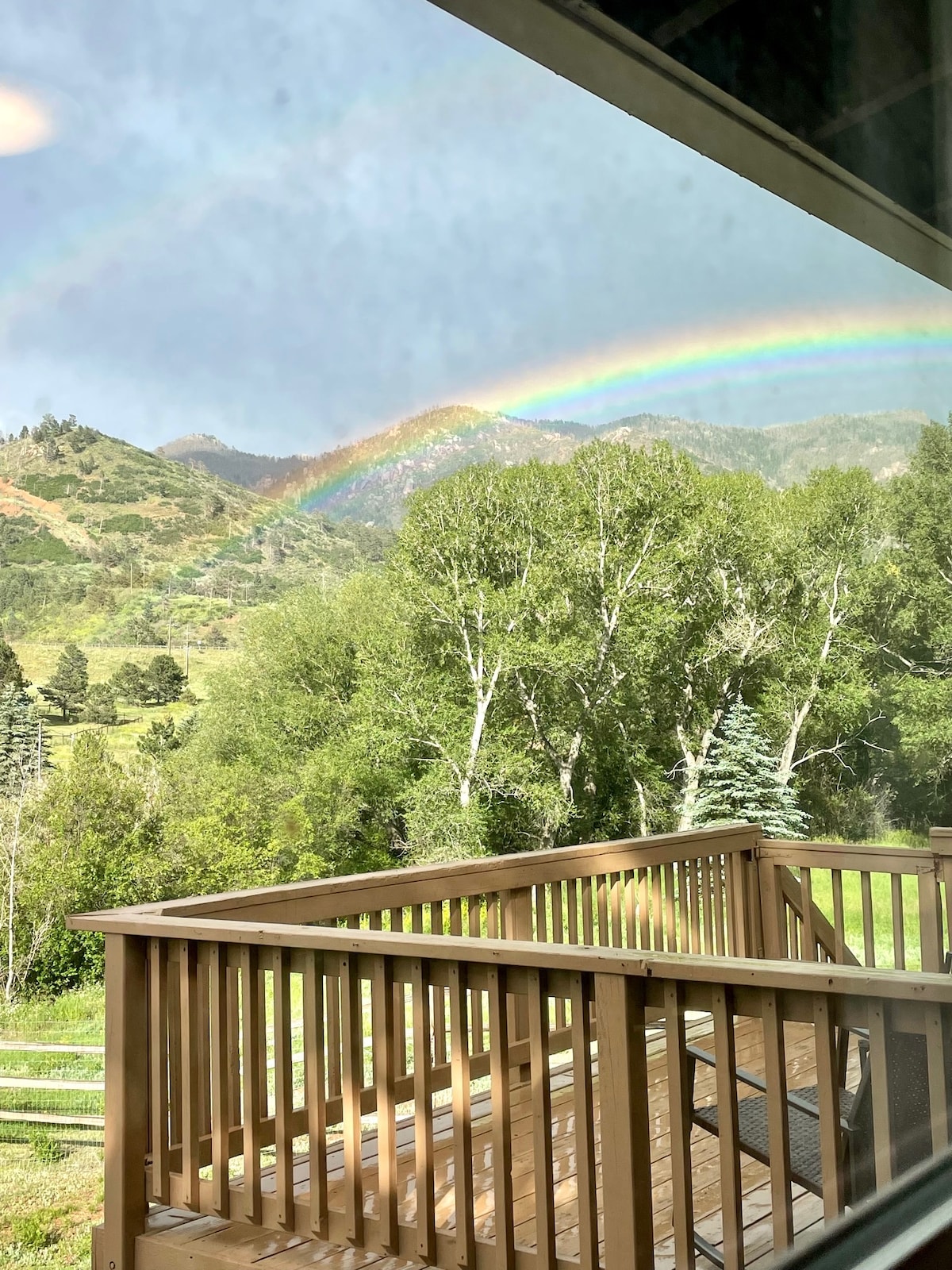 A wooden deck is viewed overlooking vibrant greenery and rolling hills. A rainbow arcs gracefully across the sky, lending color to the mountainous backdrop. Lush trees frame the scene, and a peaceful atmosphere is evident in the surrounding nature.