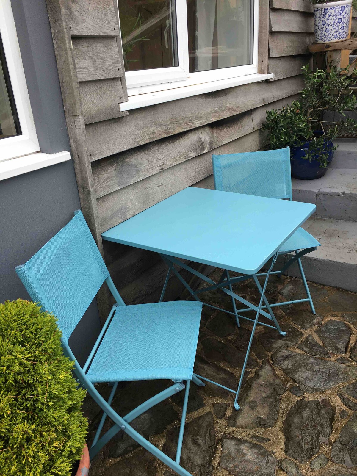 A small outdoor seating area features a blue metal table with two matching folding chairs. The table is set against a wooden exterior, with a stone path leading to a few steps. Potted greenery adds a touch of nature to the setting.