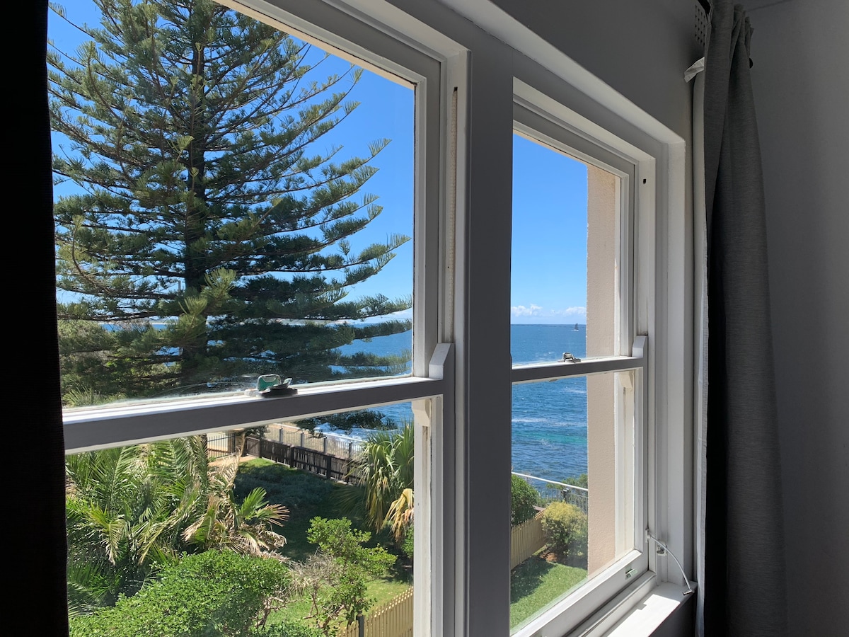 Large windows frame a view featuring a tall pine tree and the ocean. Bright natural light filters through, enhancing the serene atmosphere of the space. The greenery of palm plants can be seen in the foreground, providing a touch of nature.