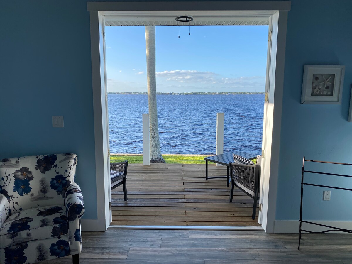 A view through an open doorway reveals a serene scene of the Caloosahatchee River. Two chairs are positioned on a wooden deck, overlooking the water. A floral-patterned chair is placed on the left side of the image, against a backdrop of soft blue walls.