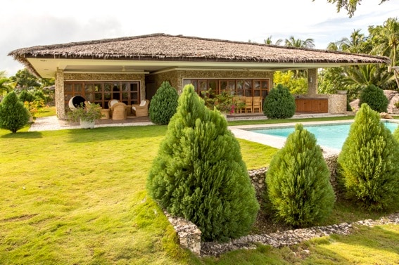 The villa is presented with a thatched roof and stone facade, surrounded by manicured greenery. A private pool features in the foreground, with landscaped shrubs framing the view. Ample windows allow natural light into the living space, enhancing the open outdoor areas.