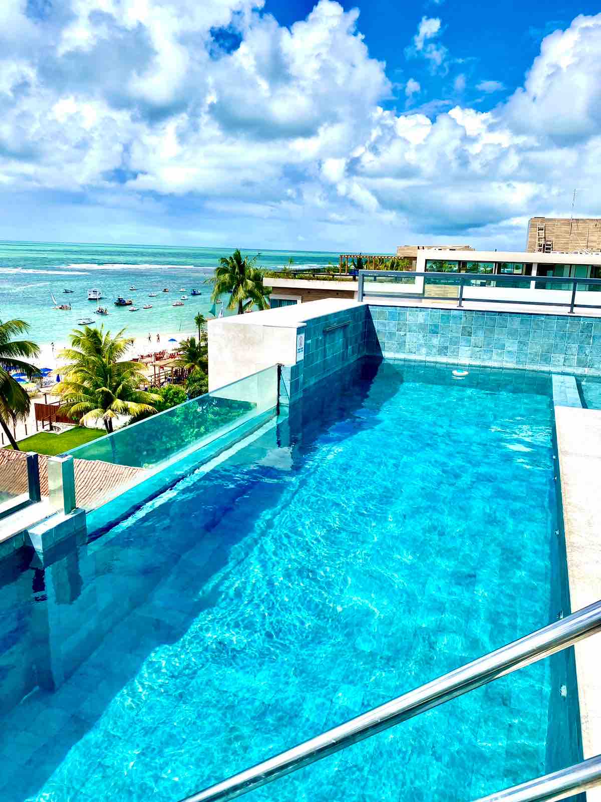 A rooftop infinity pool extends towards the horizon, merging with the vibrant blue sea. Tall palm trees line the waterfront, and boats can be seen in the distance. The bright sky is accented by scattered clouds, creating a serene coastal atmosphere.