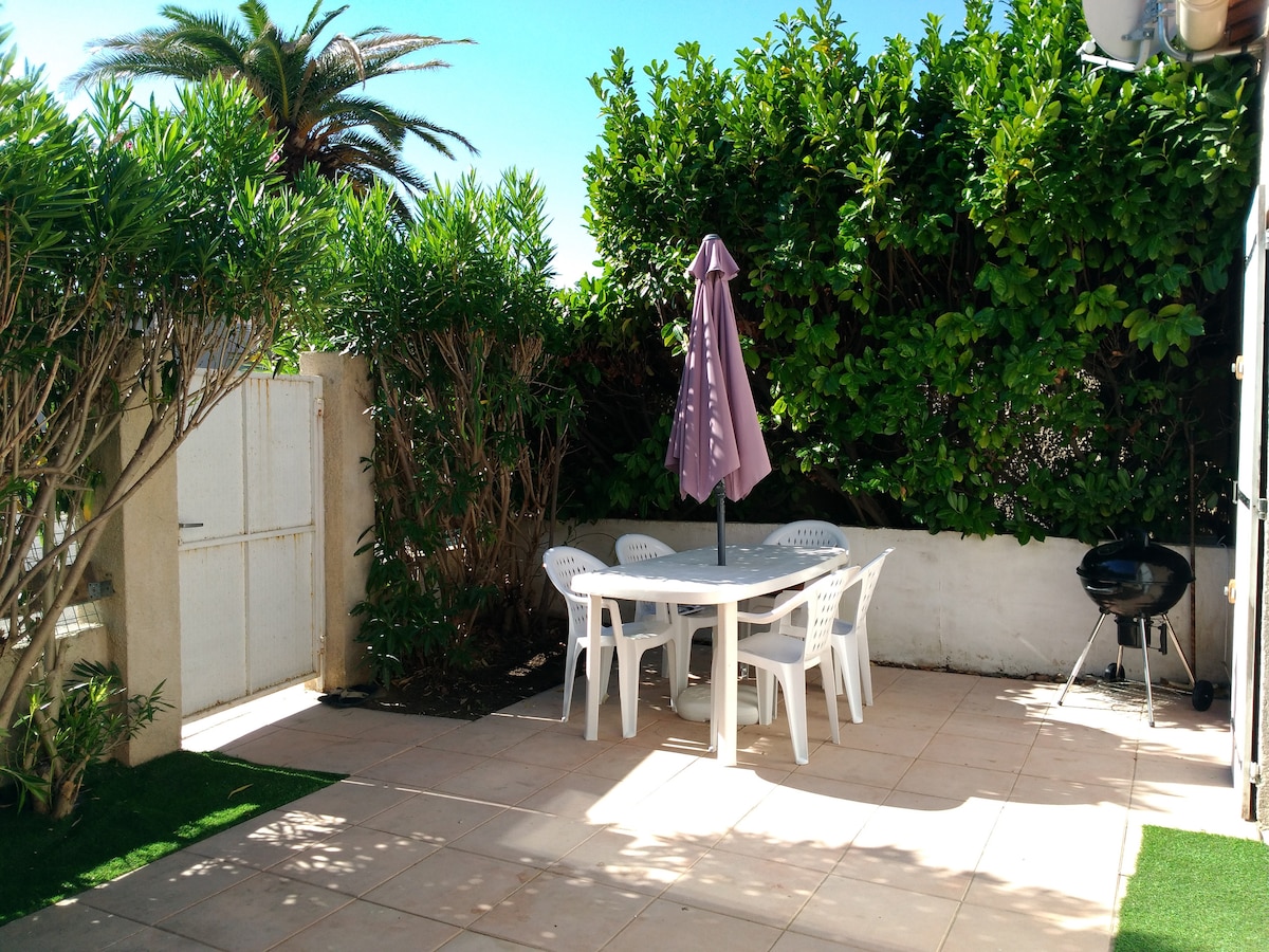 A private garden space features a round table surrounded by four white chairs, shaded by a large umbrella. Lush greenery provides privacy, while a charcoal barbecue is positioned nearby. The patio is paved with light tiles, creating a clean and open atmosphere.