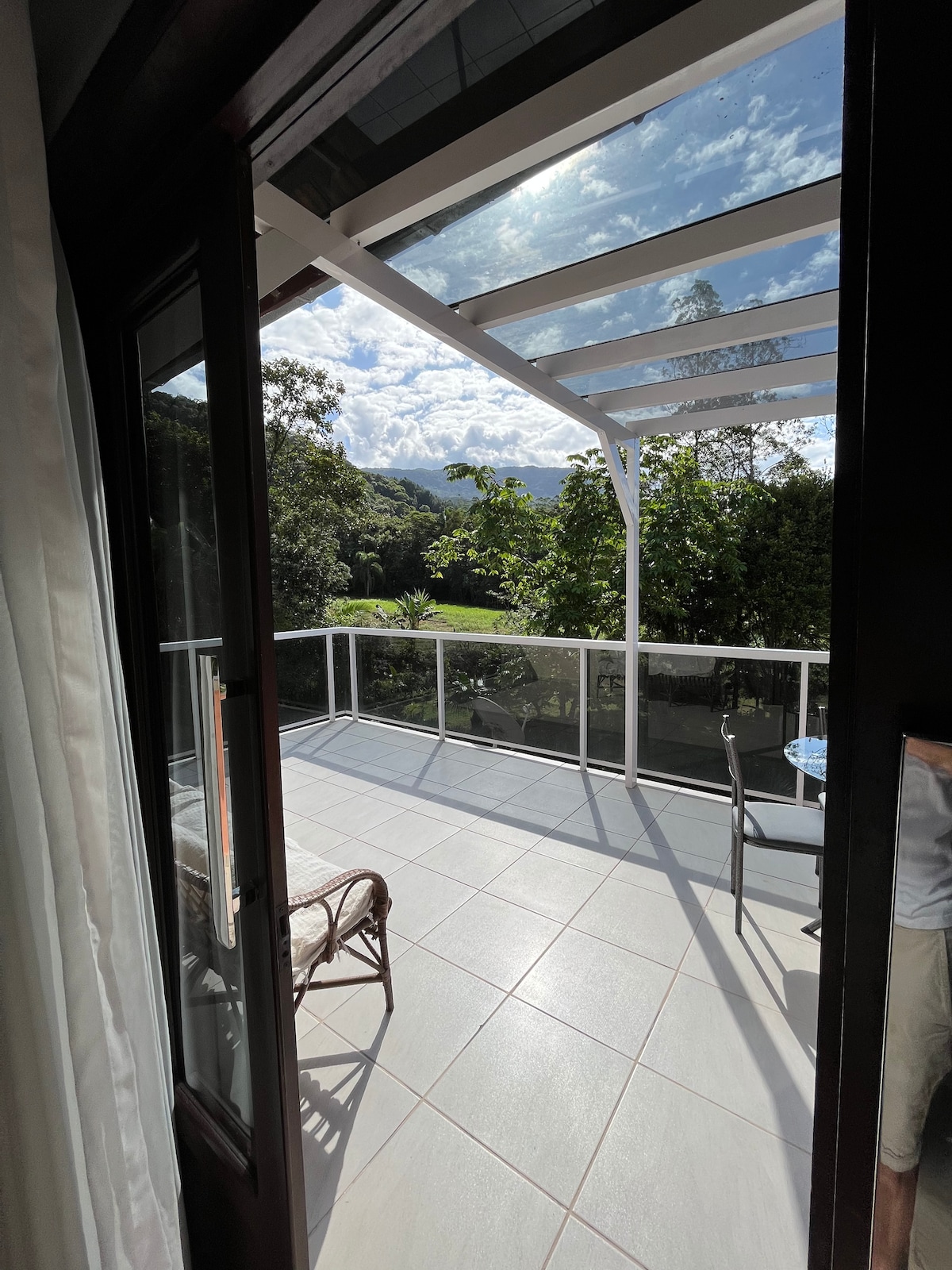 A spacious balcony is visible, featuring a glass railing that offers views of the surrounding greenery. A table and a chair are positioned near the entrance, with the sunlight casting light across the tiled floor. Lush trees and distant mountains can be seen in the backdrop.