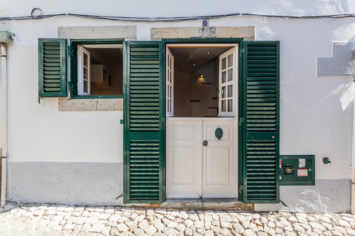 The entrance features double green shutters and a white door. The doorway opens into a well-lit interior, with hints of a kitchenette visible through the open windows. A textured stone path leads to the entrance, enhancing the charming exterior.