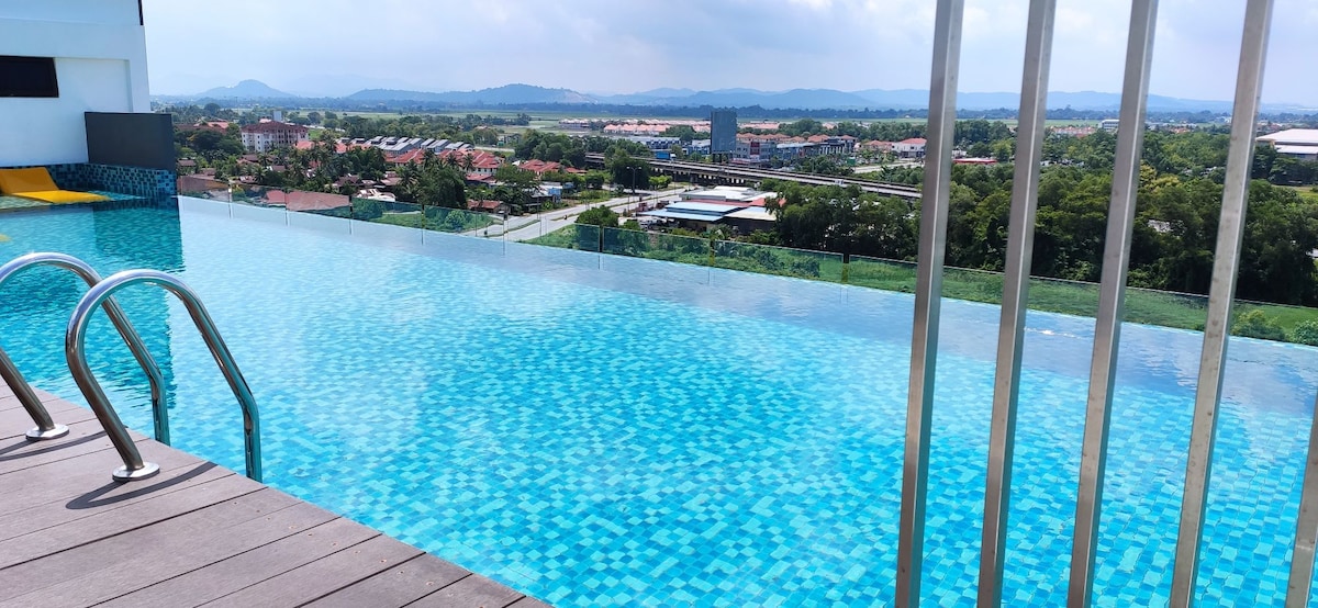 An infinity pool is framed by sleek metal railings, offering a clear view of the surrounding landscape. The water displays a mosaic of blue tiles, reflecting the sky. Lush greenery and a distant skyline are visible beyond the pool.