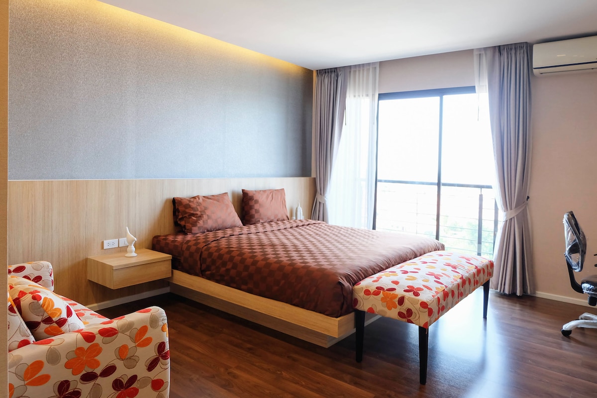 A spacious bedroom features a large bed with a patterned bedspread, accompanied by a floral upholstered bench. Natural light enters through a large window, creating a bright atmosphere. A comfortable chair and a work desk are visible, enhancing the room's functionality.