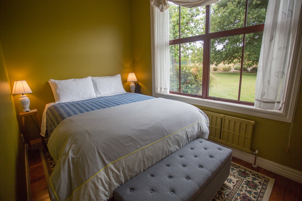 A comfortable queen bed is positioned in front of a large window, framed by light curtains. Soft lamps provide gentle illumination beside the bed. A plush ottoman sits at the foot of the bed, with a view of the green garden visible through the window.