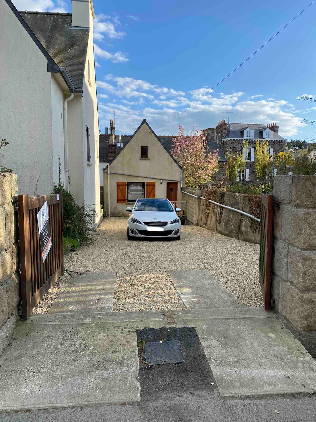 A gravel driveway leads to a charming house with a sloped roof, framed by greenery on either side. A vehicle is parked at the entrance, with blue skies and wispy clouds visible in the background, indicating a bright day.