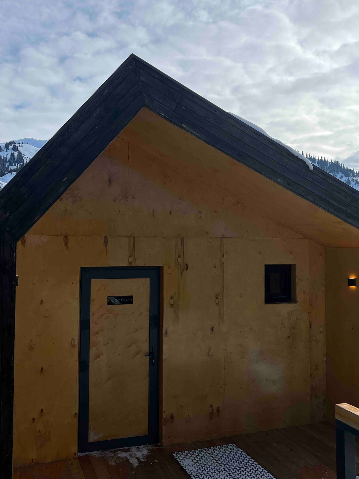 The exterior of a modern cottage is showcased, featuring a sloped roof and wooden walls. A single door and a small window are visible, with a light fixture on the side. The backdrop includes hints of snowy mountains under a cloudy sky.