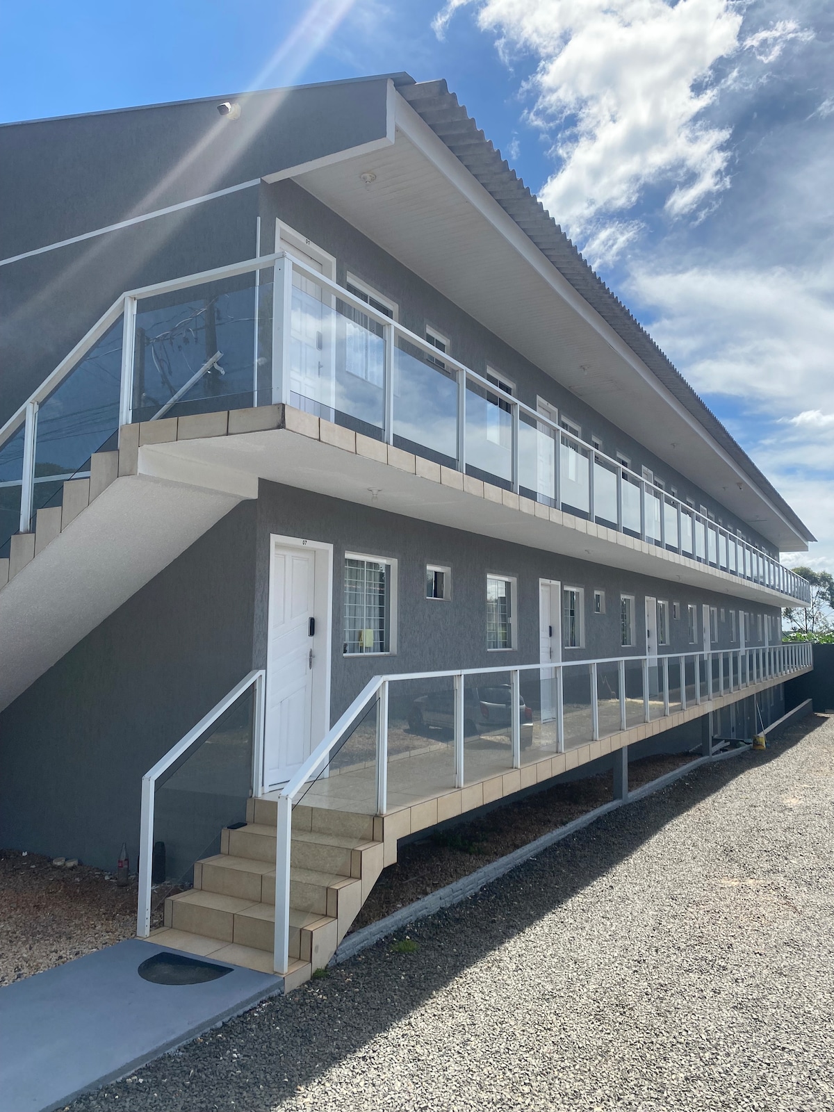 A multilevel building is presented with a modern design. The exterior features a sleek gray facade and glass railings along the balcony. Stairs provide access to the upper level, while a smooth walkway leads to individual unit doors. Sunlight brightens the scene under a partly cloudy sky.
