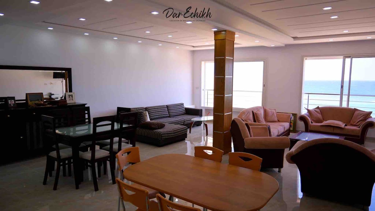 The spacious living area features a sectional sofa and armchairs arranged around a coffee table. A dining table accommodates six, with natural light flooding in through large windows that offer views of the sea. Elegant lighting fixtures are installed in the ceiling.