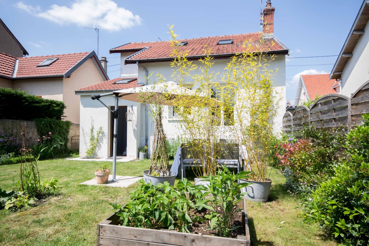A serene garden space is featured, complete with a spacious lawn and various greenery. A patio area is marked by a sunshade and comfortable seating. Raised garden beds are visible, showcasing plants. The charming house with large windows sits in the background, bathed in natural light.