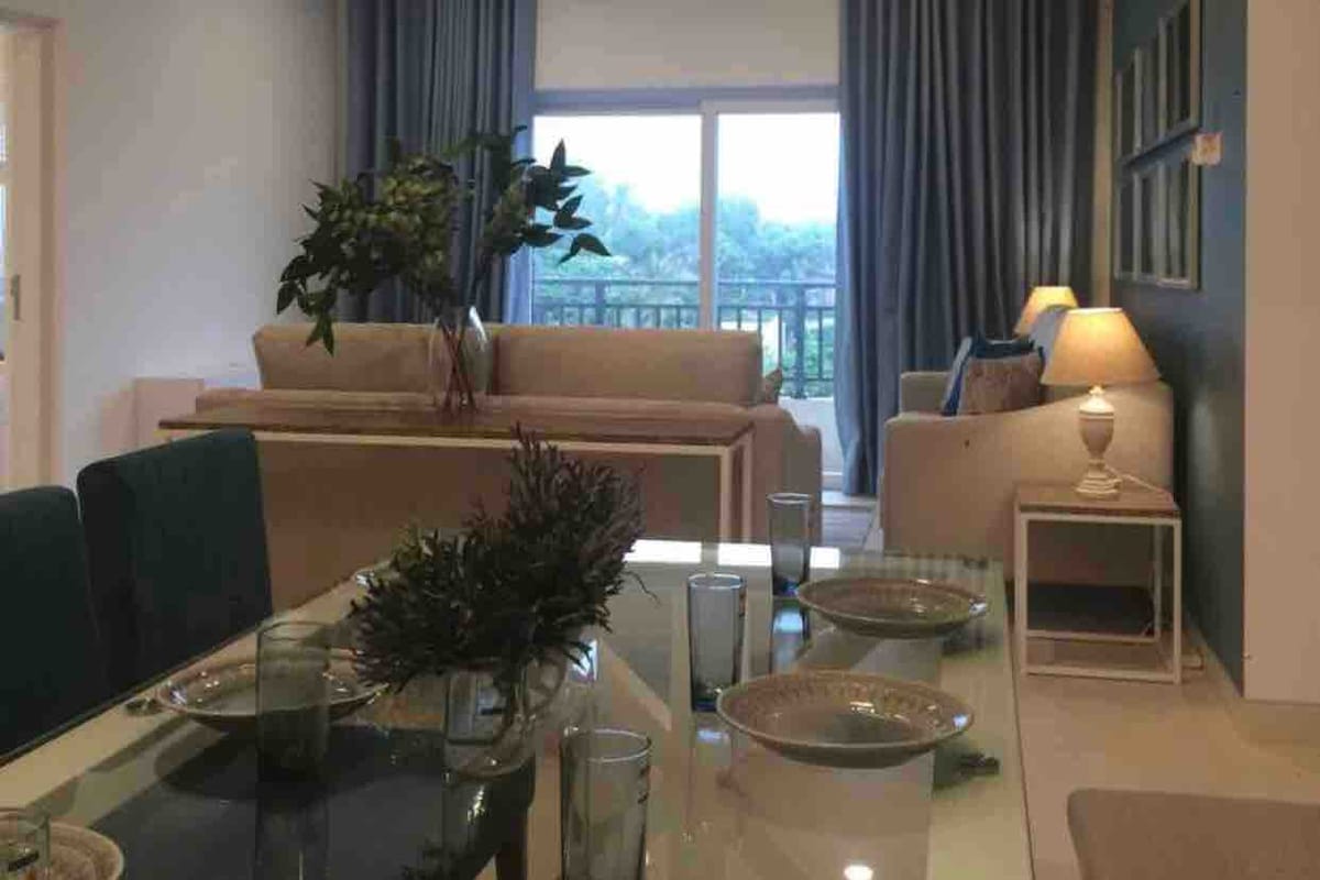 A bright living area showcases a spacious couch and armchair, complemented by soft blue curtains. A dining table is set with plates and glasses, while a vase filled with greenery adds a touch of freshness. Natural light streams through large sliding doors, leading to a balcony.