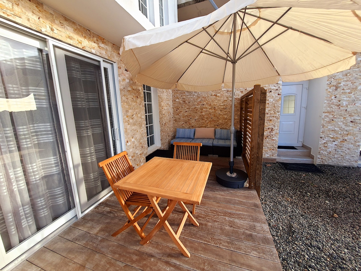 An outdoor seating area features a wooden table and two folding chairs, positioned on a wooden deck. A large, cream-colored umbrella provides shade over a cushioned lounge space. Light-colored stone walls surround the area, enhancing the inviting ambiance.