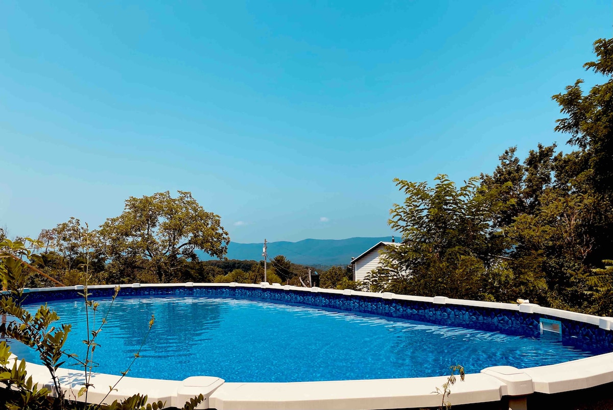 A tranquil pool is visible against a clear blue sky, edged by a well-defined white railing. Lush greenery surrounds the area, enhancing the view of distant mountains and offering a serene atmosphere for relaxation.