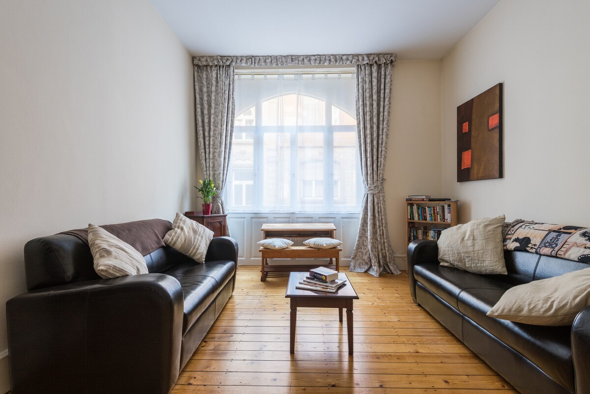 A spacious sitting area features two comfortable sofas facing each other, with decorative pillows adding subtle texture. A small wooden coffee table sits between them, and a large window allows natural light to fill the room, framed by elegant curtains. A bookcase is positioned nearby.
