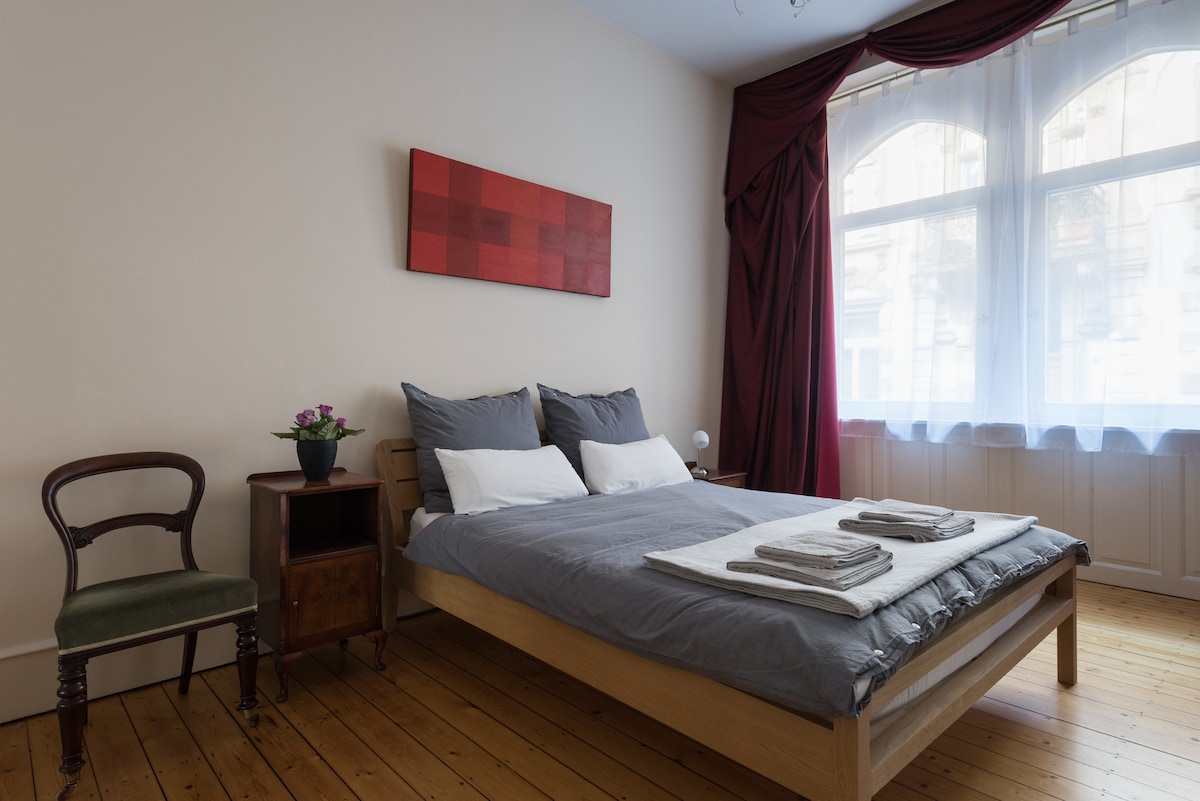 A spacious bedroom features a comfortable bed dressed in soft gray linens. Natural light filters through large windows, complemented by elegant drapes. A small bedside table holds a decorative plant, and a wooden chair is positioned nearby, enhancing the room’s simplicity and functionality.