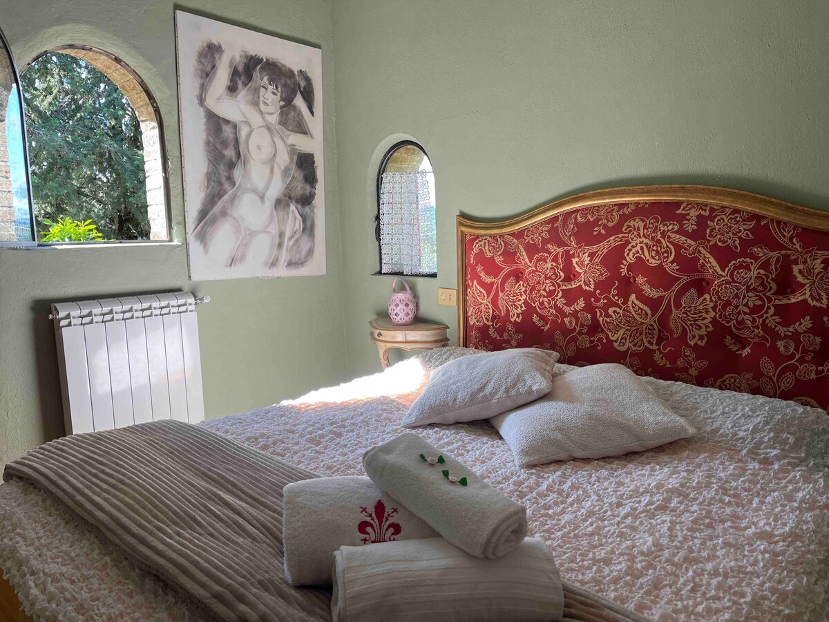 A bedroom features a spacious bed with decorative bedding and multiple pillows. A piece of art hangs on the wall above the headboard. Natural light enters through two arched windows, framing views of greenery outside. A radiator is placed beside the bed.