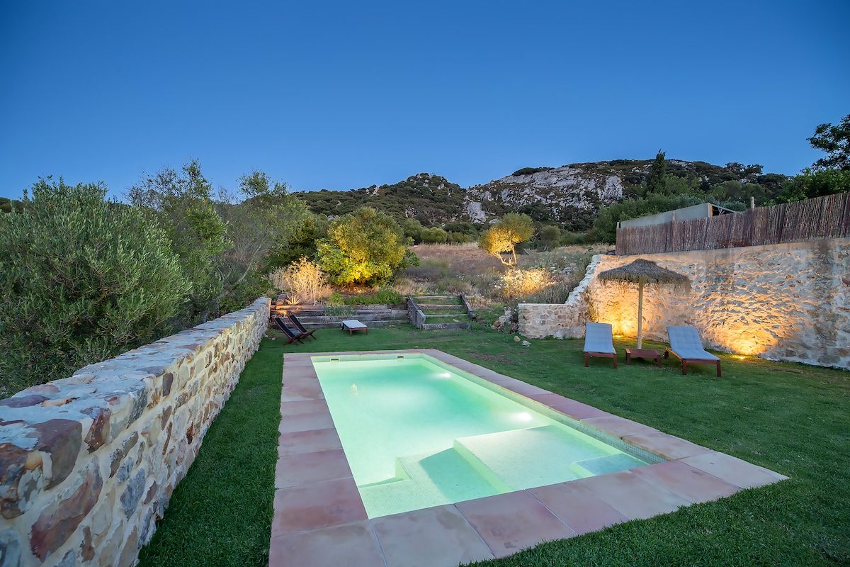 A serene pool area is surrounded by lush greenery and natural stone walls. Two loungers provide a comfortable space for relaxation. Soft lighting highlights the pool, inviting a peaceful atmosphere as twilight descends over the landscape.