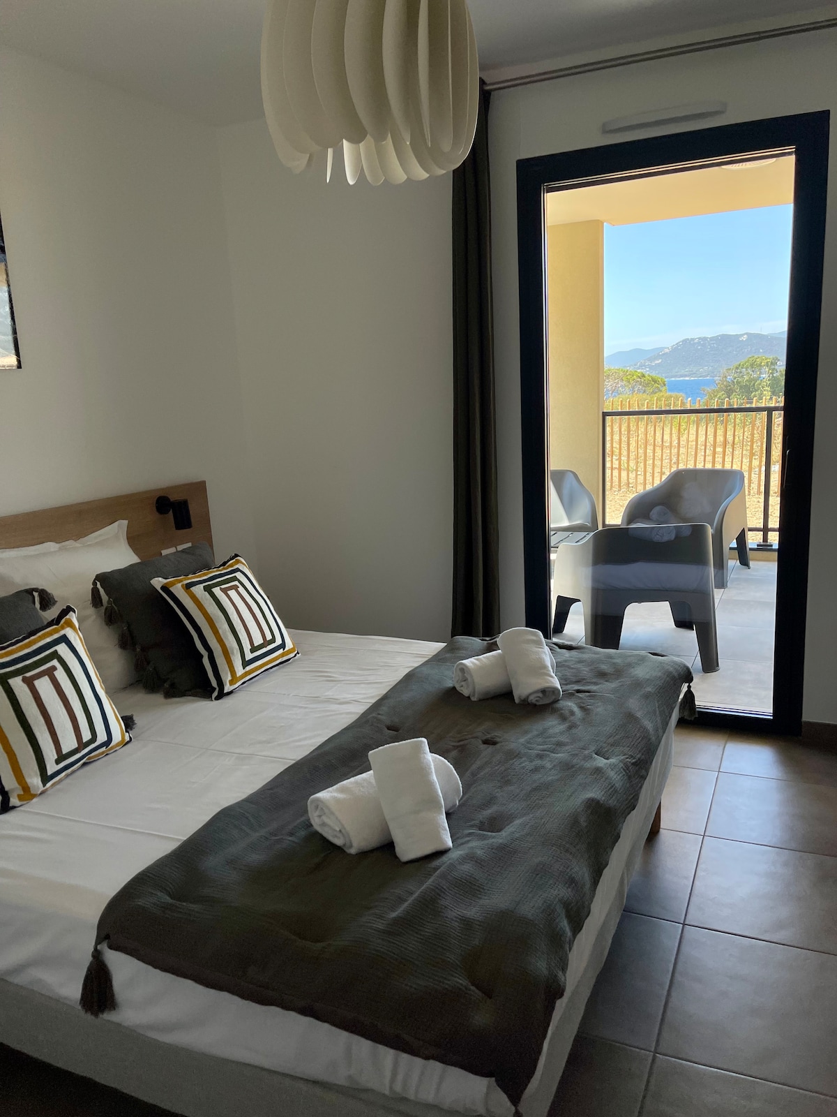 A well-appointed bedroom features a spacious bed dressed with a dark throw and neatly arranged white towels. Light streams in through sliding glass doors, which lead to a balcony. Decorative pillows add a modern touch to the inviting atmosphere.