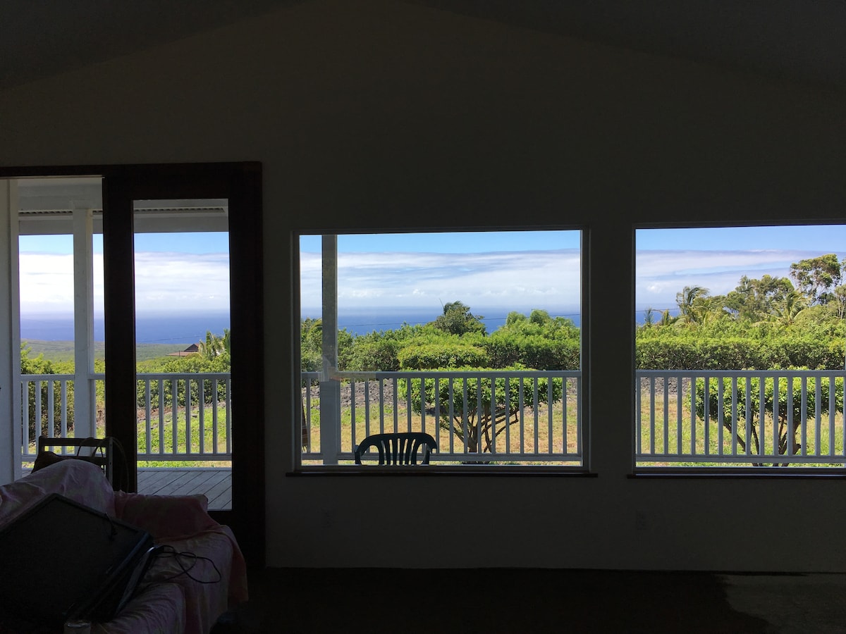 Large windows frame sweeping ocean views, allowing natural light to fill the open space. The gentle landscape includes greenery visible through the glass, enhancing the serene atmosphere. A simple chair is positioned nearby, complementing the inviting setting.