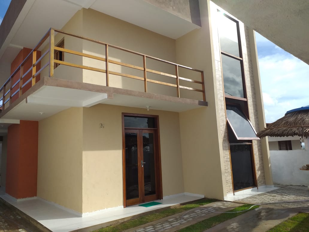 The exterior of the property features a modern design with a light-colored facade and large glass windows. A balcony is visible on the upper level, providing an open space for relaxation. The entrance is marked by a wooden door, with a pathway leading to the front yard.
