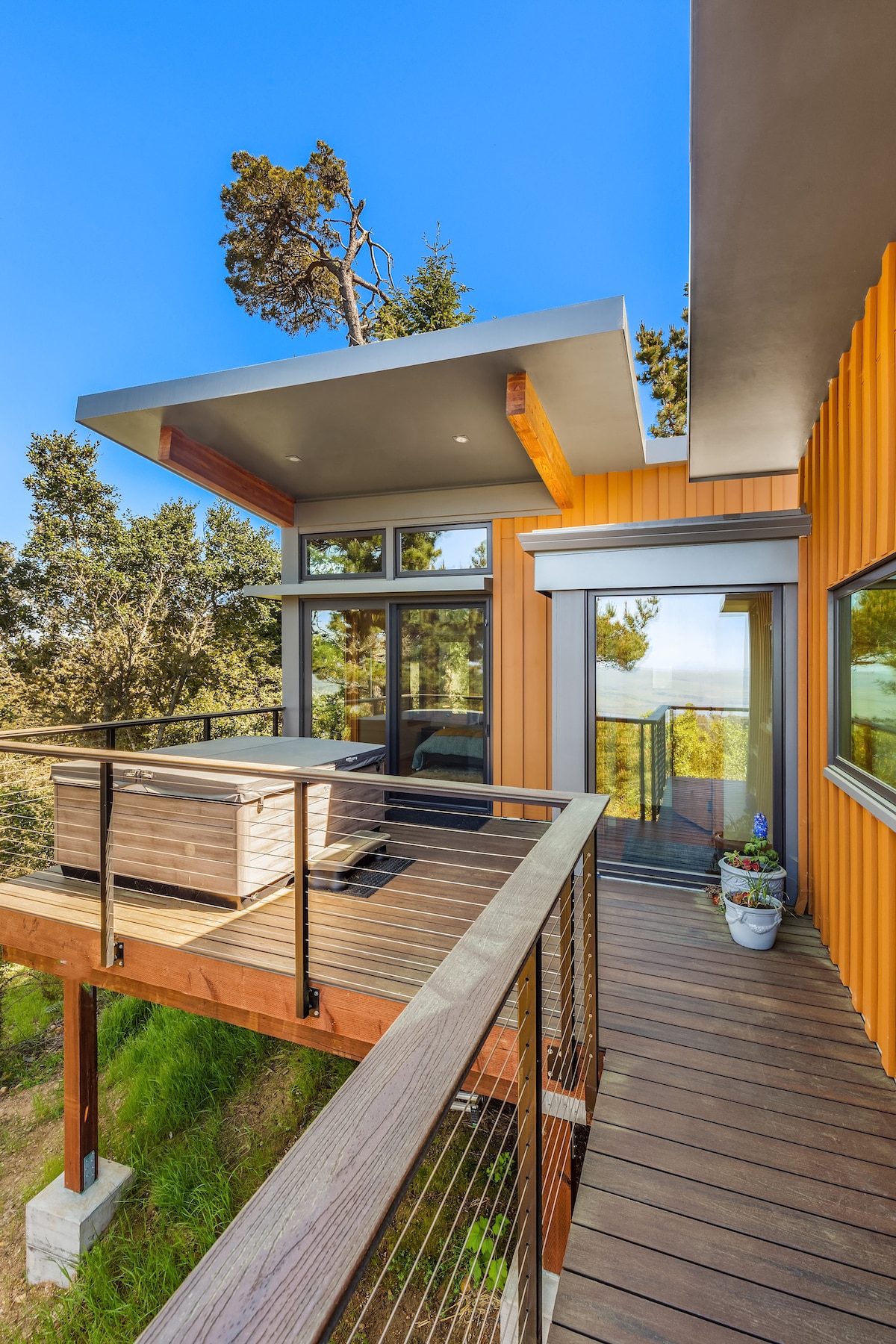 A contemporary exterior view showcases a well-designed deck surrounding the house. The orange siding contrasts with the greenery beyond, while large windows and a covered area provide ample natural light. A hot tub is visible on the deck, surrounded by potted plants.