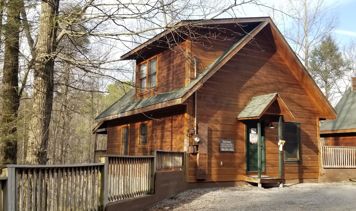 The cabin's exterior is constructed of rich wooden siding, blending harmoniously with the surrounding forest. A welcoming entrance features a green door, complemented by matching shutters. Gravel pathways lead to a spacious porch, accentuated by rustic railings and a natural wooded backdrop.