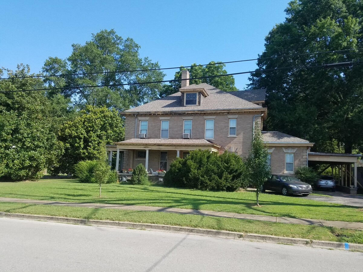 A historic two-story home with a stone facade is surrounded by greenery. A wrap-around porch is visible, with several chairs placed on it. The lawn is well-maintained, and a car is parked on the side. Tall trees provide shade in the area.