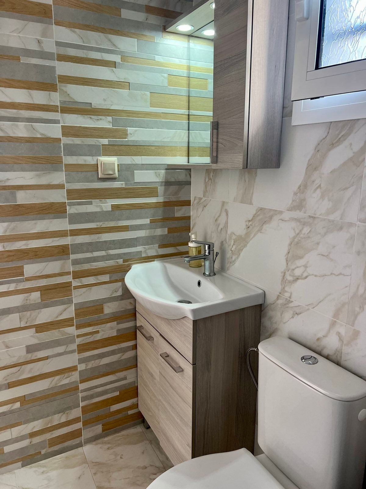 A contemporary bathroom features a compact sink unit with wooden cabinetry and modern fixtures. The walls are adorned with a mix of tiles in neutral and earthy tones, complemented by a large mirror that reflects natural light. The toilet is positioned efficiently within the space.