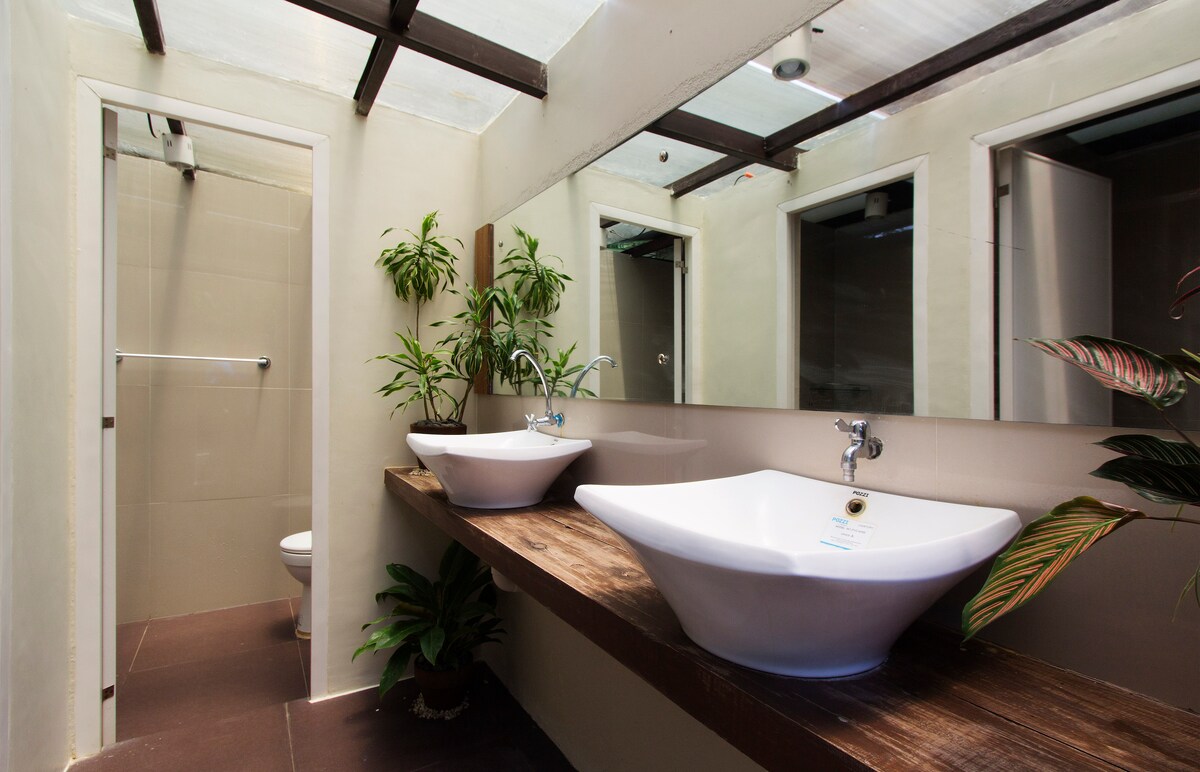An airy bathroom features dual sink basins on a wooden countertop, surrounded by natural plants. A glass shower area is visible, along with a separate toilet stall. Mirrors reflect the bright lighting from above, enhancing the open feel of the space.