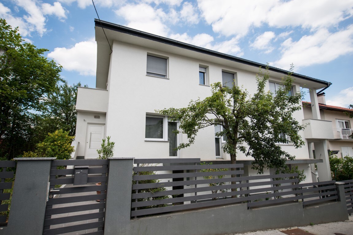 The exterior of a modern, multi-story building is presented, characterized by a clean white façade and large windows. Green trees and shrubs are visible in the foreground, providing a natural contrast against the structure. A gray fence outlines the property, ensuring privacy.