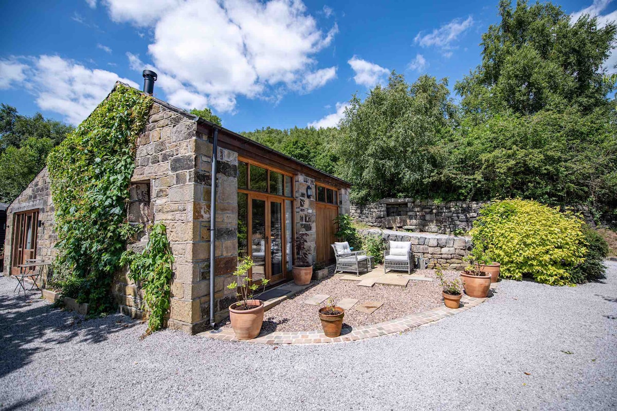 An inviting barn exterior is framed by lush greenery and sunny skies. The stone façade is complemented by large windows, allowing natural light to fill the interior. Potted plants add a touch of warmth to the gravel pathway leading to comfortable outdoor seating.