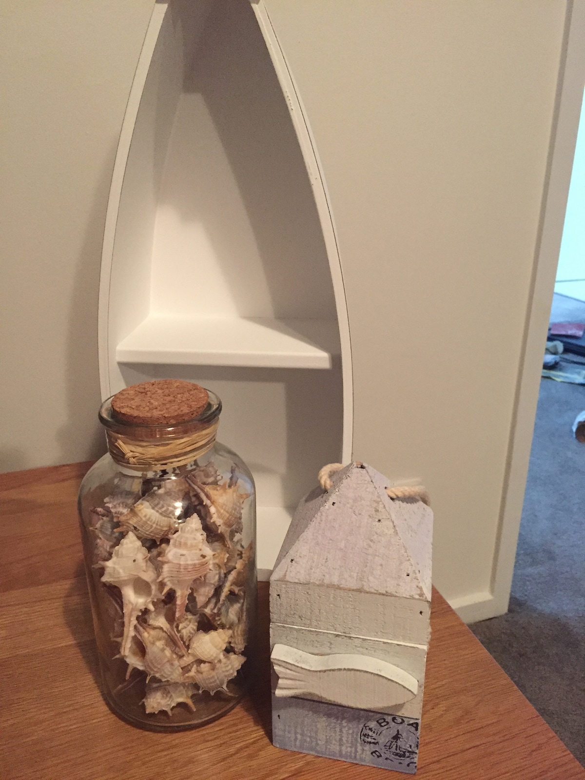 A wooden boat-shaped shelf is displayed against a neutral wall, with a glass bottle filled with seashells and a decorative wooden house nearby. The bottle features a cork stopper, while the house showcases soft pastel colors and textured details.