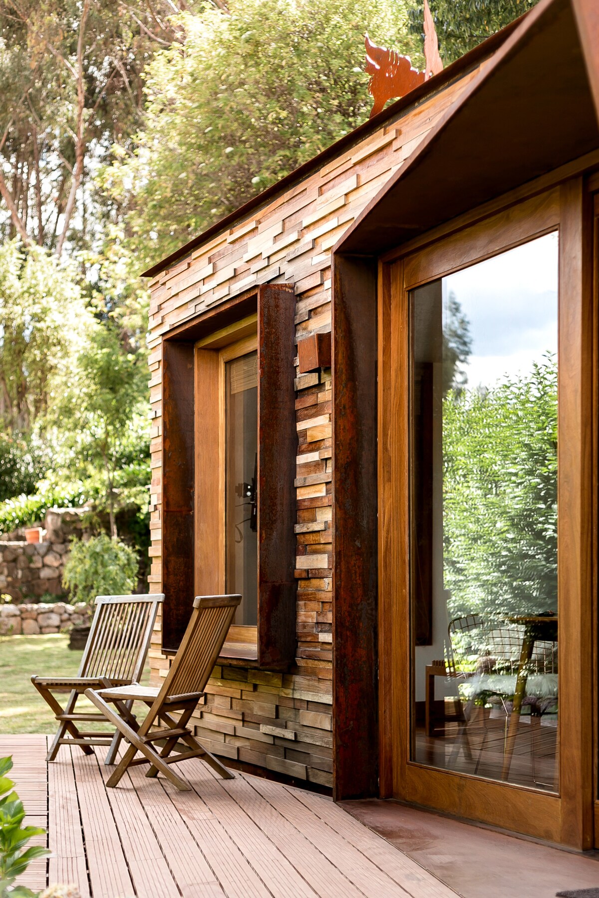 A cabin exterior showcases a unique stone and wood facade, with two wooden chairs positioned on a spacious deck. Large glass doors provide a view of the lush greenery, enhancing the connection with the natural surroundings.