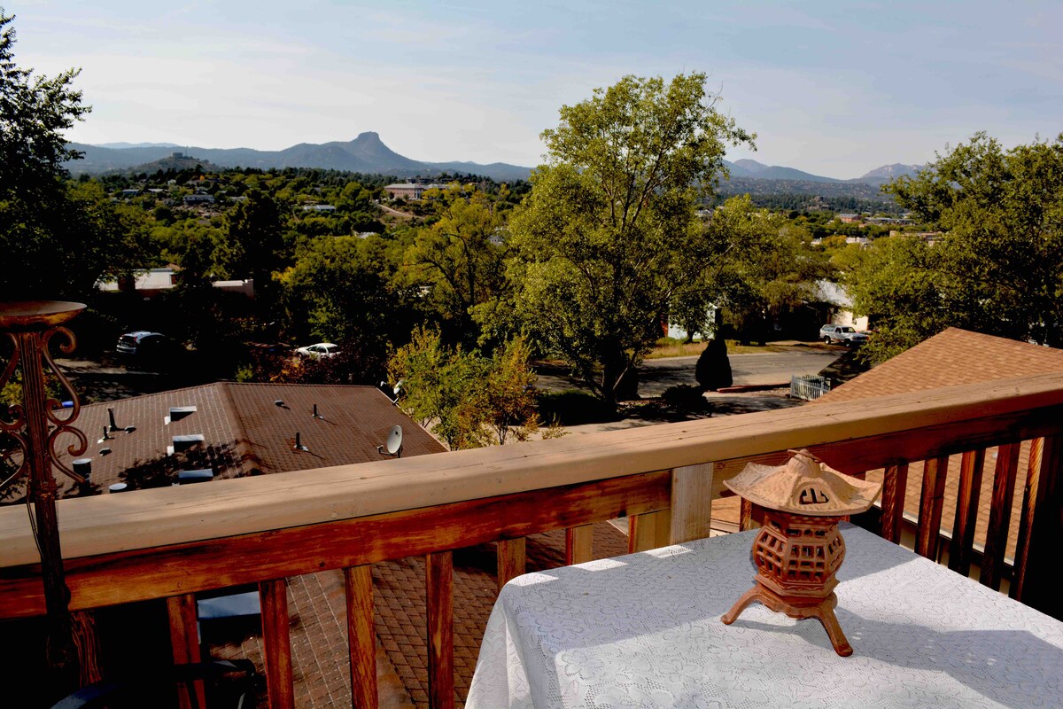 A scenic view from a balcony features rolling hills and distant mountains under a clear sky. A decorative lantern sits on a white tablecloth, and trees provide a natural frame to the landscape, enhancing the sense of tranquility.