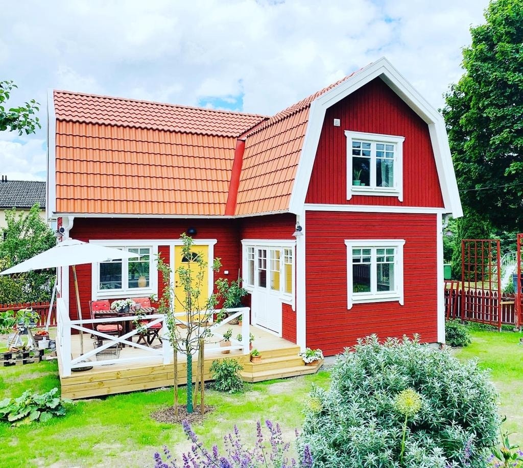 A charming red house with a pitched roof is surrounded by well-maintained gardens. A wooden deck area features outdoor seating beneath a large umbrella, with flower beds and greenery framing the property. Large windows allow for ample natural light, adding to the inviting exterior.