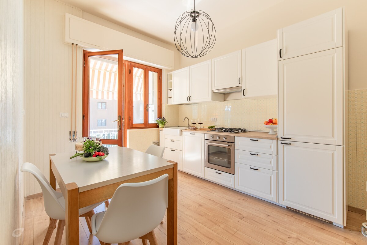 A modern kitchen is featured with white cabinetry and stainless steel appliances. A wooden dining table accommodates four and is set with fresh fruit. Natural light filters in through large windows, enhancing the airy feel of the space.