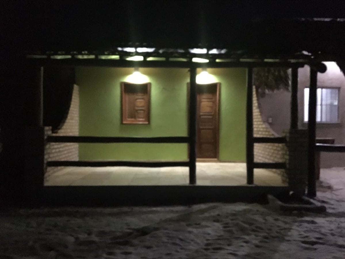 A night view of the vacation home is presented, featuring a porch with two wooden doors. Soft lighting illuminates the entrance, highlighting the green walls. The surrounding sandy area adds to the serene setting near the beach.