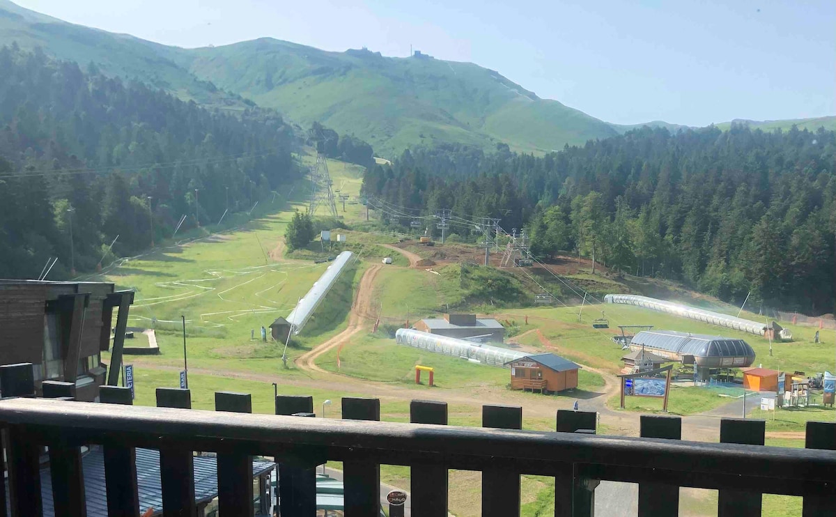 A scenic view of verdant hills and a ski area is captured from the balcony. Elevated ski lifts and installation structures are visible in the foreground, surrounded by lush trees. The landscape reflects a mix of natural beauty and recreational facilities, promoting a serene outdoor ambiance.