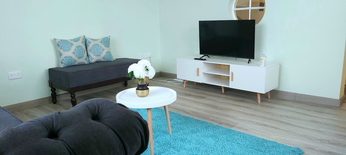 The living area features a light blue accent wall, a comfortable sofa adorned with patterned cushions, and a sleek white entertainment unit with a television. A round coffee table sits on a soft area rug, complemented by a decorative vase of flowers.