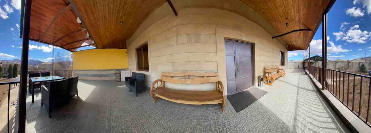 A spacious outdoor area features a wooden bench against a textured stone wall, complemented by a dark seating arrangement nearby. A set of double doors invites entry, while large windows allow natural light to illuminate the space.