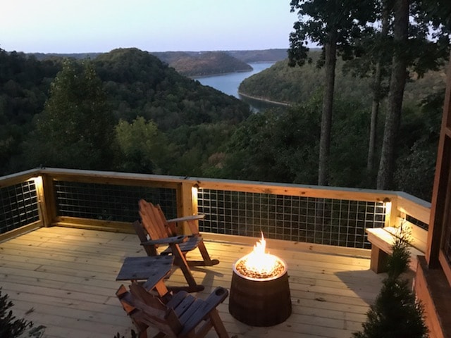 An outdoor deck features a cozy seating area with two wooden chairs facing a fire pit. The surrounding trees provide a natural setting, and the view of the lake and rolling hills is visible in the distance as daylight fades.