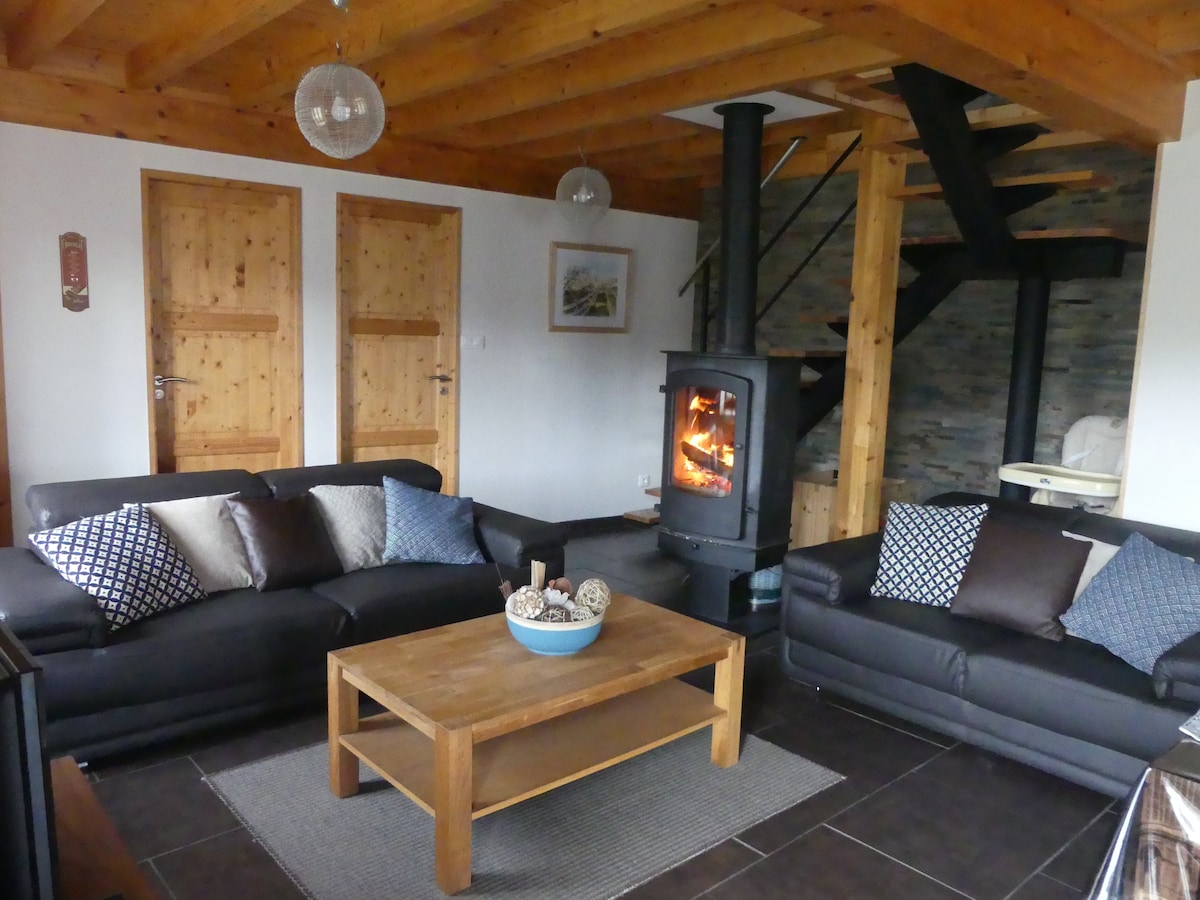 A cozy living area features two dark leather sofas facing a central wooden coffee table. A decorative bowl with natural elements rests on the table. A modern wood stove stands against a stone wall, adding warmth to the space, while a spiral staircase leads to the upper floor.