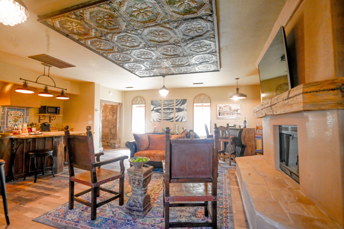 A spacious living area is centered around a stone fireplace, with wooden chairs and a plush sofa positioned for seating. Artistic wall decor complements the rich texture of the ceiling, while warm lighting enhances the inviting atmosphere. A dining area is visible in the background.