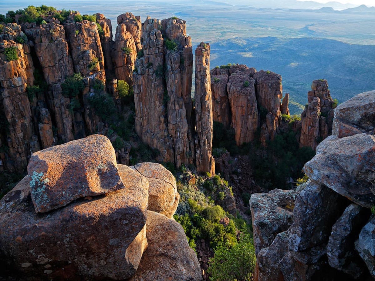 Jagged rock formations rise sharply from a rocky landscape, casting shadows over the valleys below. Lush greenery is visible along the cliffs, and a vast expanse of land stretches into the background, revealing rolling hills under a clear sky.