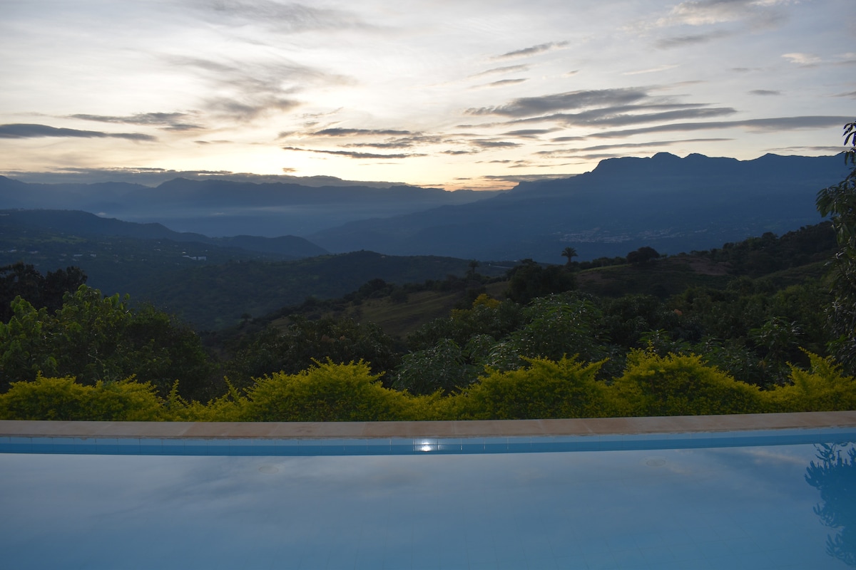 A serene landscape features distant mountains under a soft, colorful sky at sunset. A tranquil swimming pool reflects the surrounding nature and sky, bordered by lush greenery, creating a peaceful atmosphere.